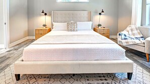 Un grand lit avec une tête de lit capitonnée, deux tables de chevet avec des lampes, une chaise recouverte d’une couverture à carreaux et un tableau encadré accroché au mur.