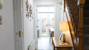 A hallway with a wooden staircase, a door with a brass handle, a table with a lamp, and a colorful rug.