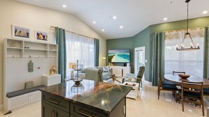 A modern living room with a kitchen area, a dining table, and a large TV screen.