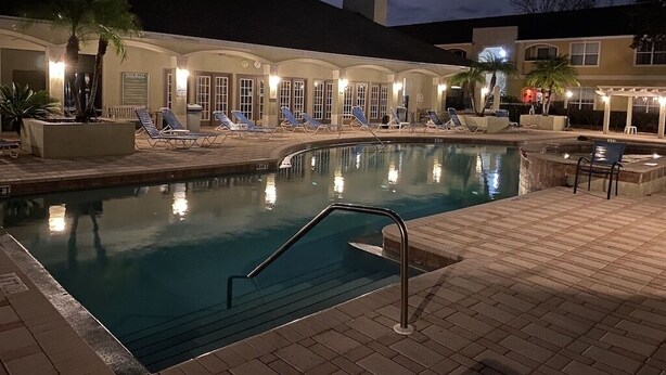 A swimming pool with lounge chairs and a covered seating area, surrounded by buildings with arched windows and a palm tree.