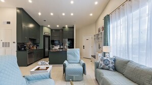 A modern living room with a sofa and armchairs, a kitchen in the background, and a large window with curtains.