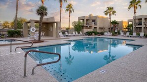 A swimming pool with lounge chairs and palm trees, surrounded by buildings.