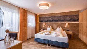 A modern hotel room with a wooden floor, a large bed with white linens, a wooden headboard, a bedside table with a lamp, and a large window with sheer curtains. - Hotel Glockenstuhl (Gerlos)