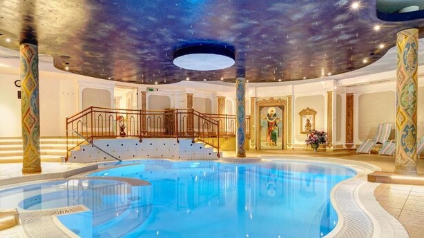 A luxurious indoor swimming pool with a starry ceiling, ornate columns, and a grand staircase leading to a balcony. - Hotel Glockenstuhl (Gerlos)