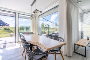 A modern dining area with a large wooden table, black chairs, and a view of the outdoors through large windows. - Lovely 8-person Country House Between the Dune Pans (Den Burg)