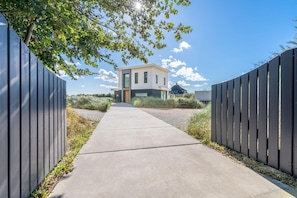 A modern building with a concrete pathway leading to it, surrounded by a metal fence and greenery. - Lovely 8-person Country House Between the Dune Pans (Den Burg)