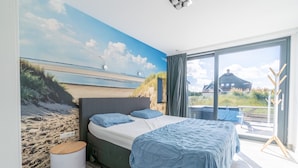 Un dormitorio con una cama grande, vista a la playa por una ventana y un balcón con mesa y sillas.