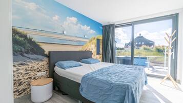 Un dormitorio con una cama grande, vista a la playa por una ventana y un balcón con mesa y sillas.