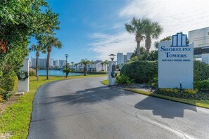 Une entrée sécurisée, fermée par un portail, donne accès aux Shoreline Towers et offre une vue dégagée sur les environs, notamment sur les palmiers et une étendue d’eau.