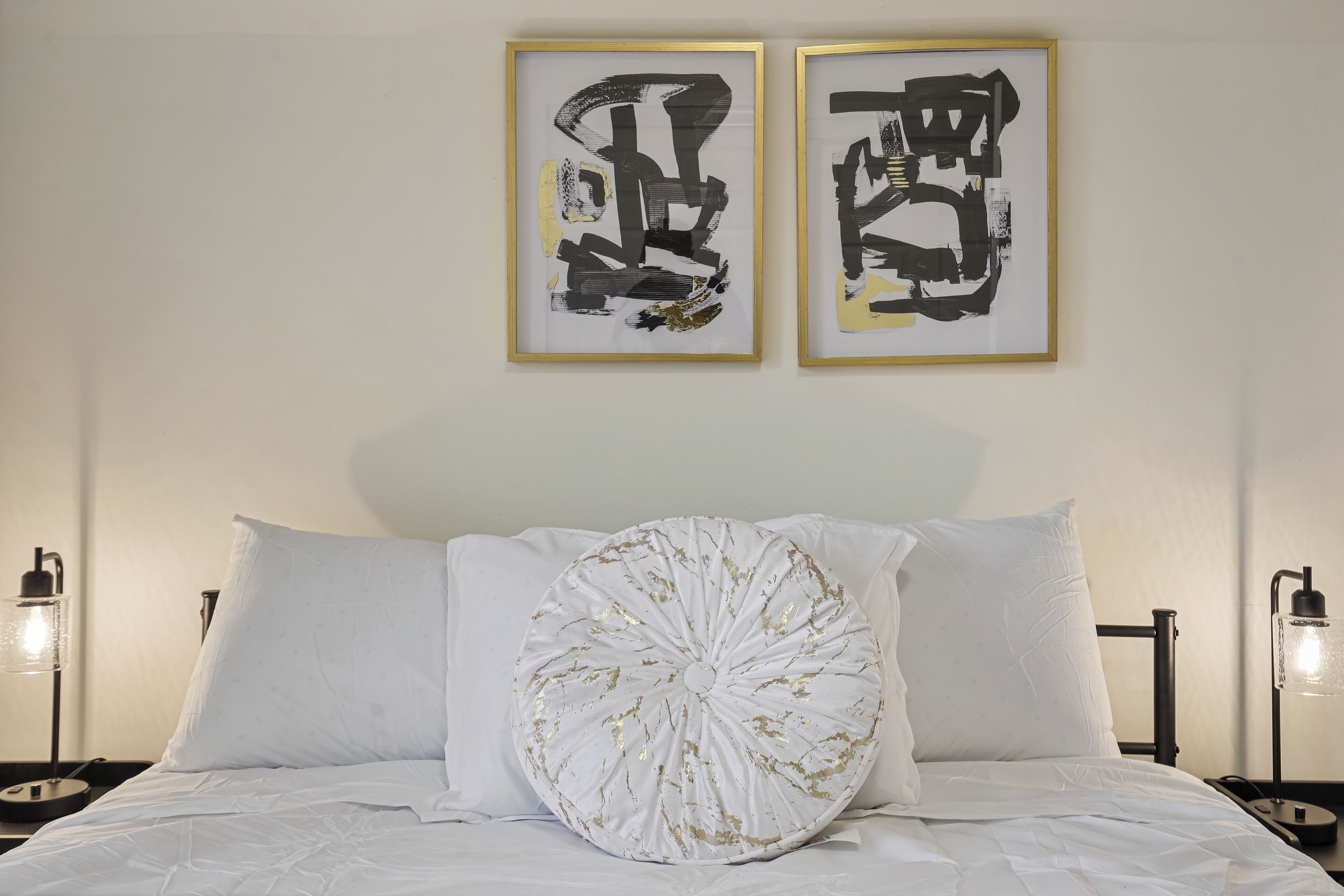 A bed with white bedding and a decorative pillow, two framed artworks on the wall, and bedside lamps.