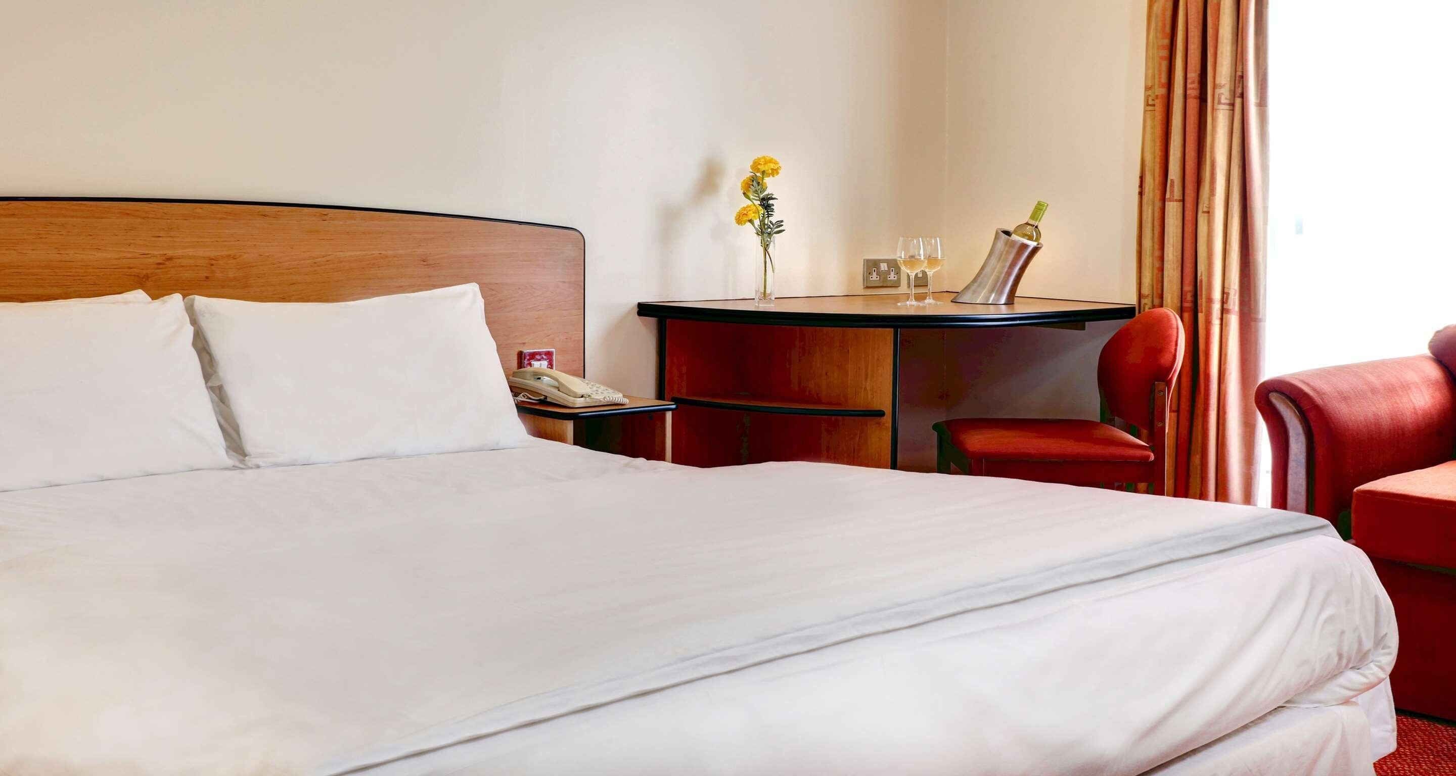 A hotel room with a large bed, a small desk with a telephone, a chair, and a table with a vase of flowers and two glasses.