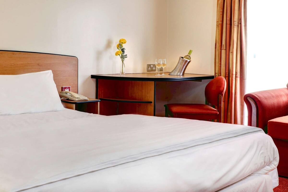Una habitación de hotel con una cama grande, un escritorio pequeño con un teléfono, una silla y una mesa con un jarrón de flores y dos vasos.