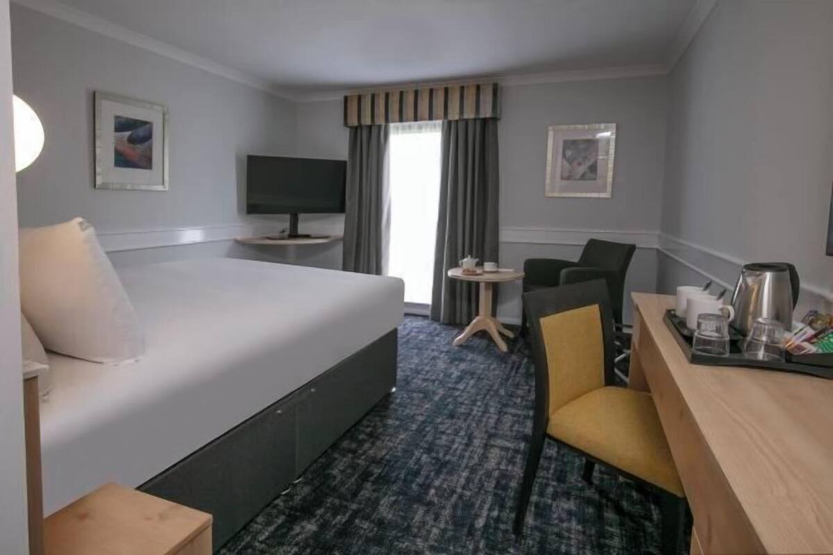 Ein Hotelzimmer mit Bett, Schreibtisch mit Tablett, Stuhl, kleinem Tisch mit Tasse und Fernseher.