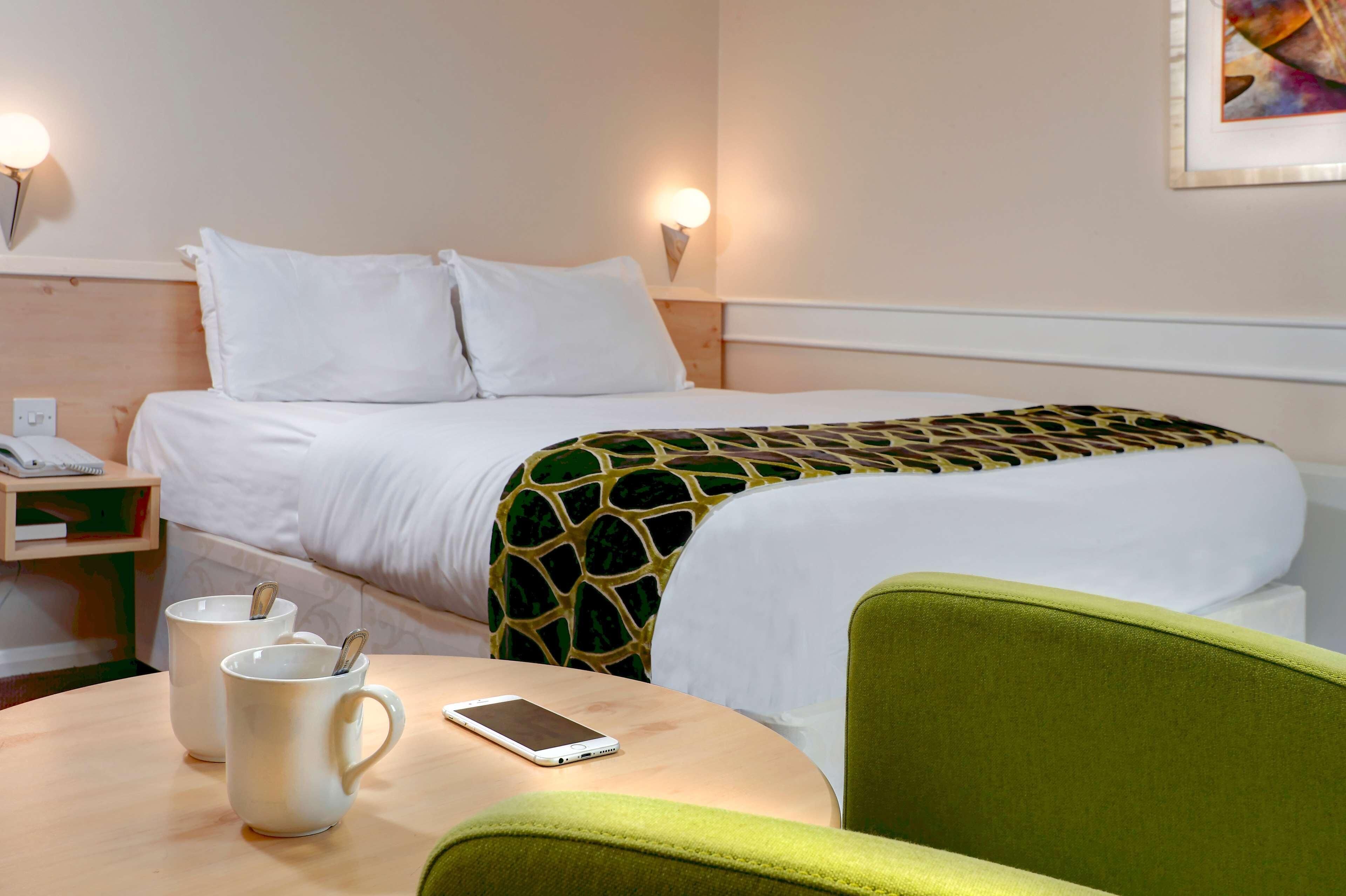 A hotel room with a bed, bedside table, two mugs, a smartphone, and a green chair.