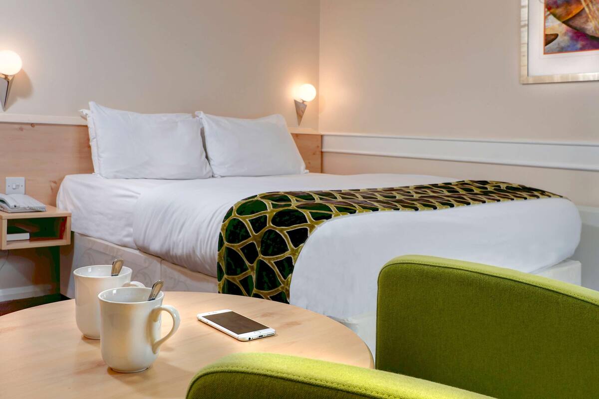 Habitación de hotel con cama, mesita de noche, dos tazas, un teléfono inteligente y una silla verde.