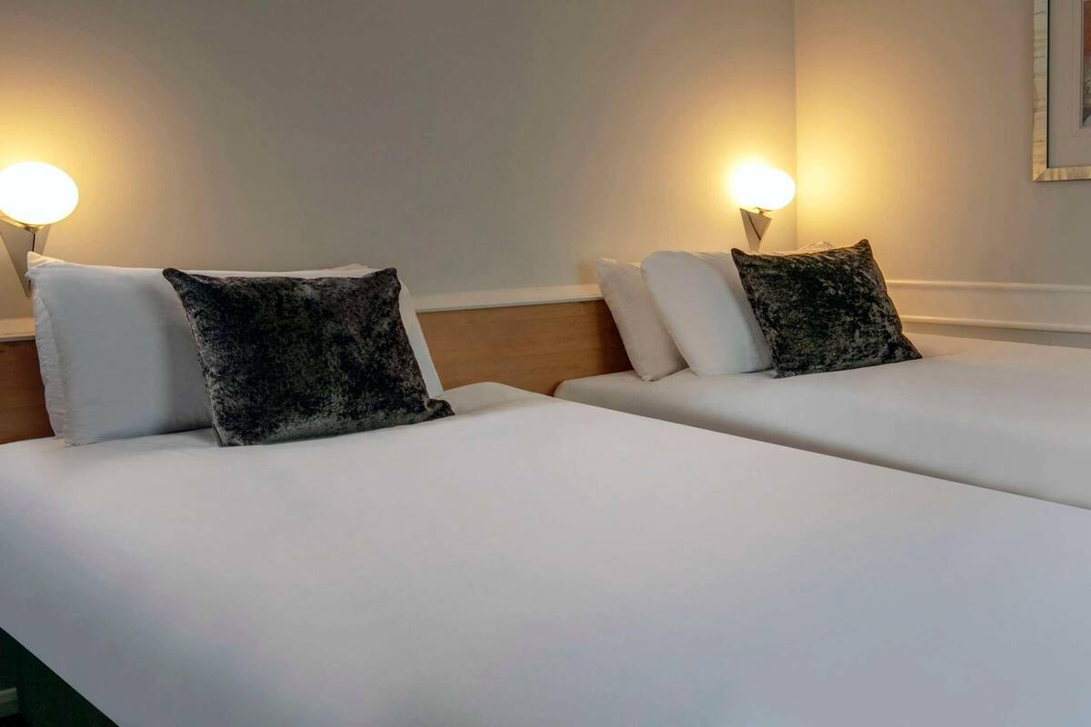 Dos camas individuales con sábanas blancas y almohadas de diseño oscuro, cada una con una luz nocturna fijada a la pared.