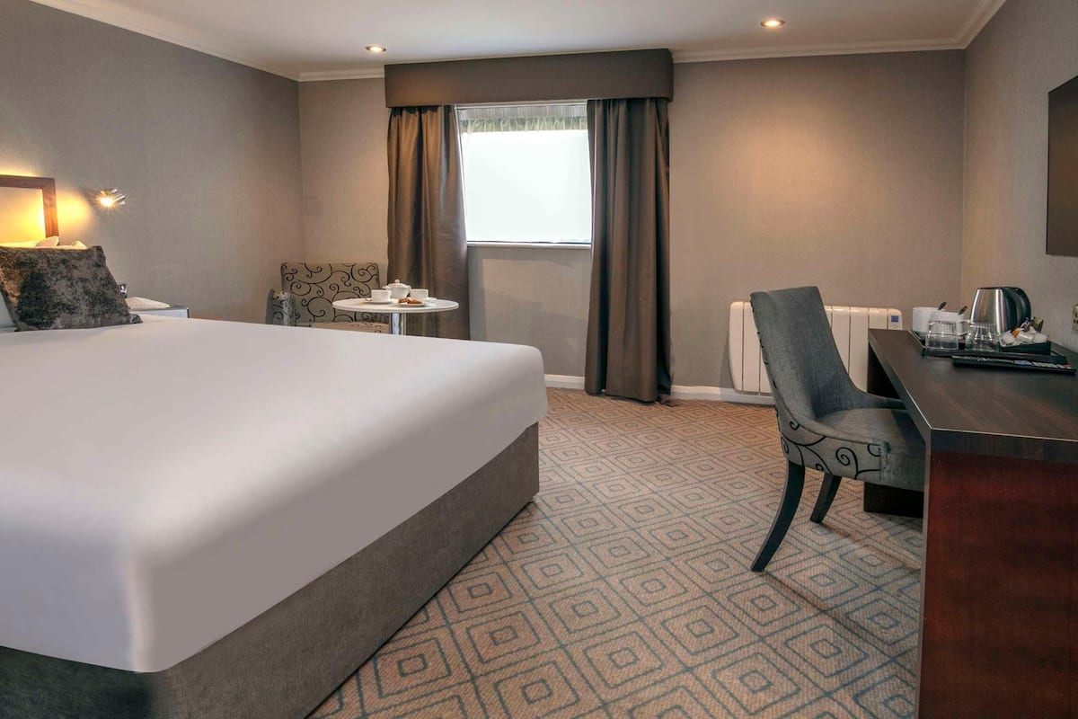 Ein Hotelzimmer mit einem großen Bett, einem Schreibtisch, einem Stuhl, einem kleinen Tisch, einem Fernseher und einem Fenster mit Vorhängen.