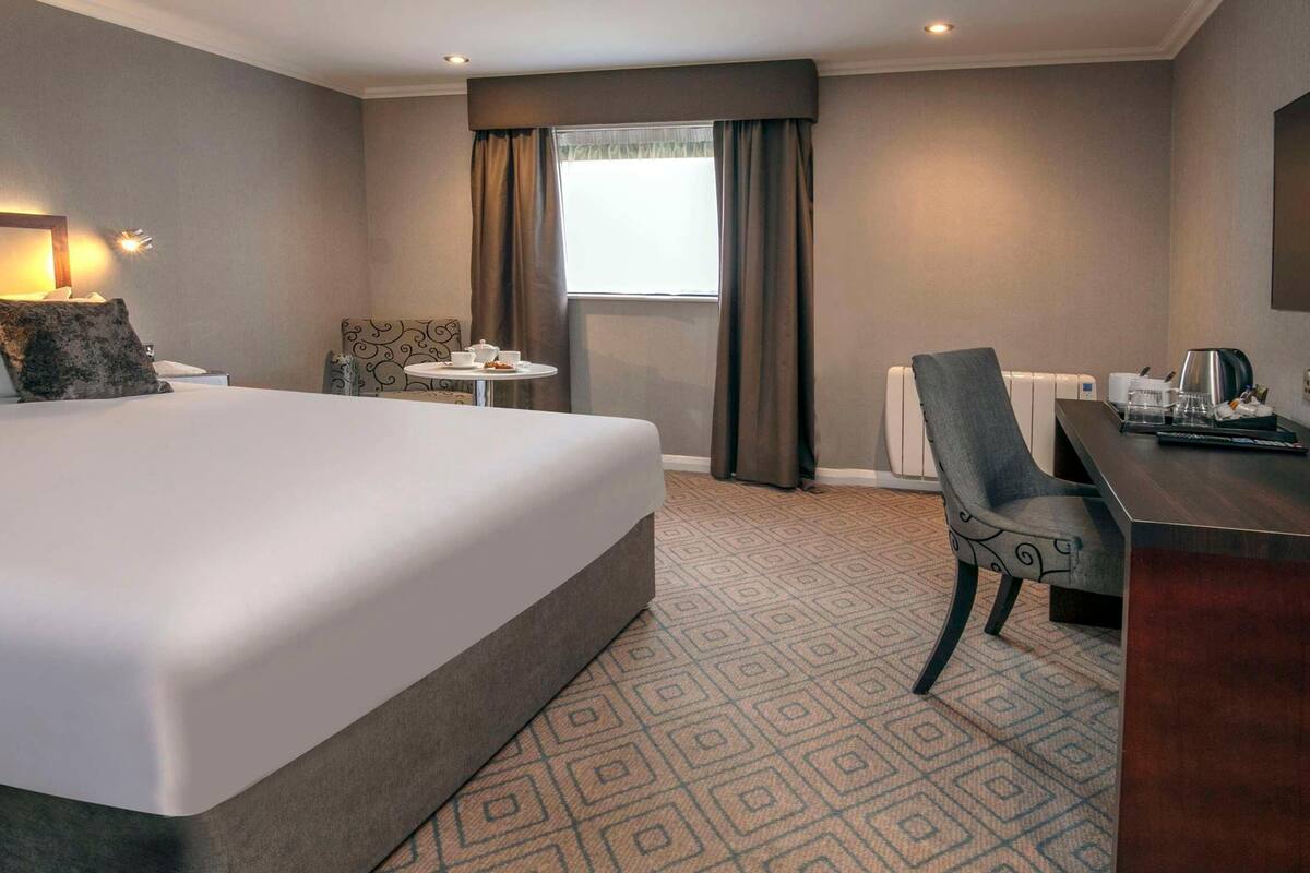 A hotel room with a large bed, a desk, a chair, a small table, a TV, and a window with curtains.