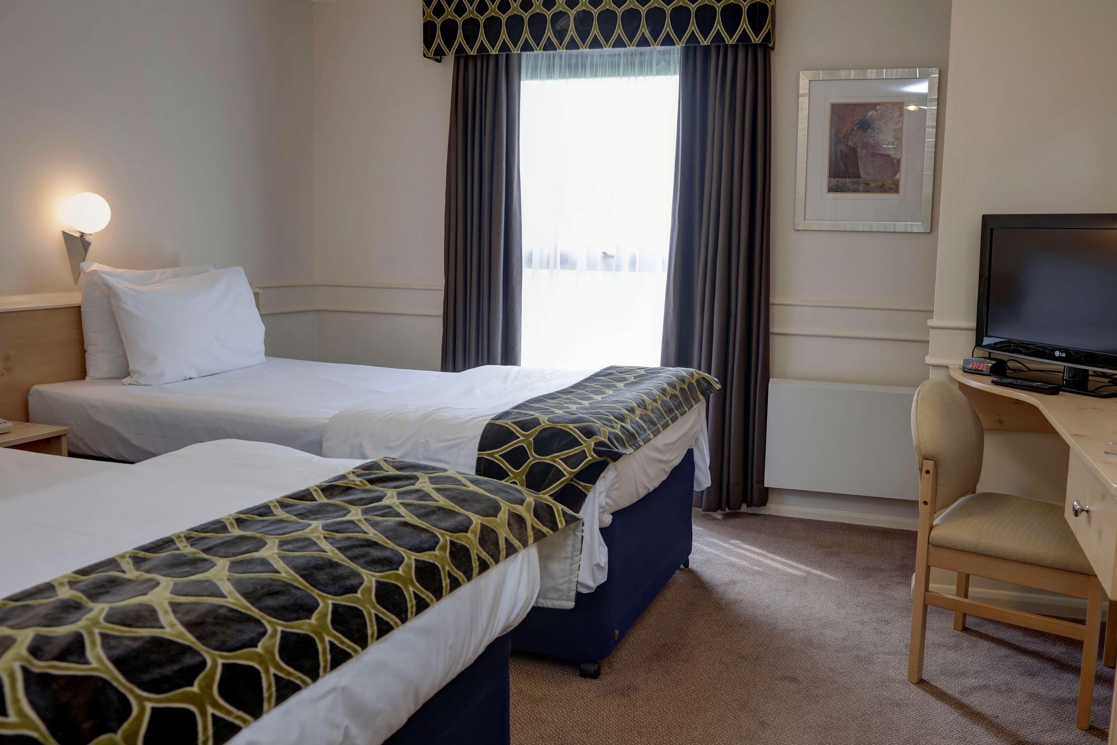 A hotel room with two beds, a desk, a chair, a television, a window with curtains, and a framed picture on the wall.