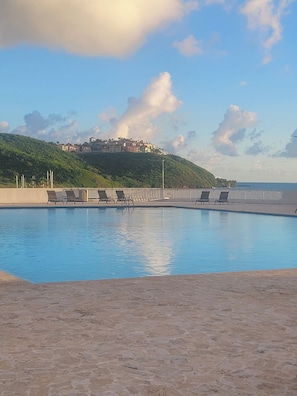 Piscina com espreguiçadeiras, vista para uma colina com edifícios e céu claro.