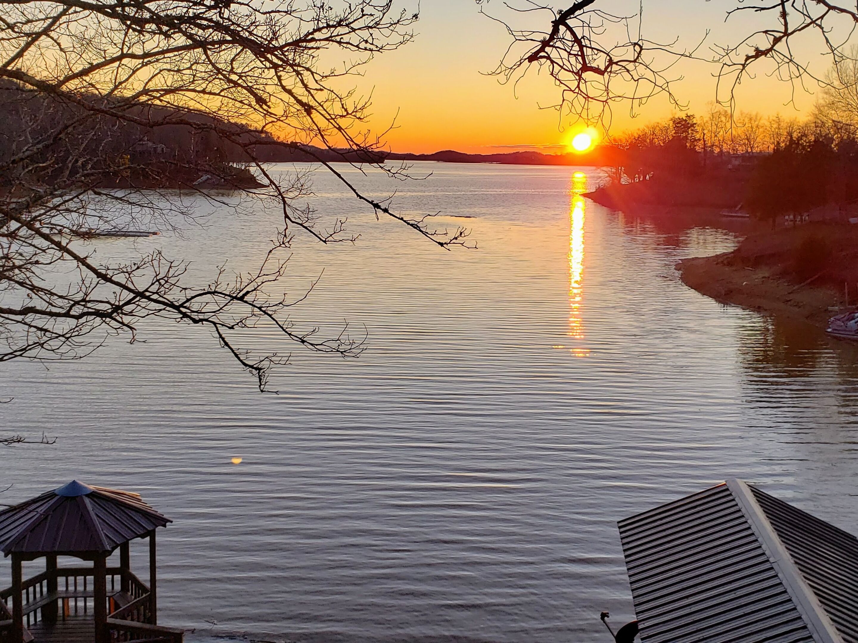 A sunset over a calm lake with a wooden dock and a gazebo.