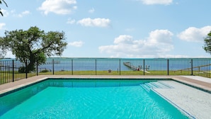 Une piscine rectangulaire à l’eau d’un bleu limpide, entourée d’une clôture en métal noir, et une zone herbeuse avec des arbres en arrière-plan.
