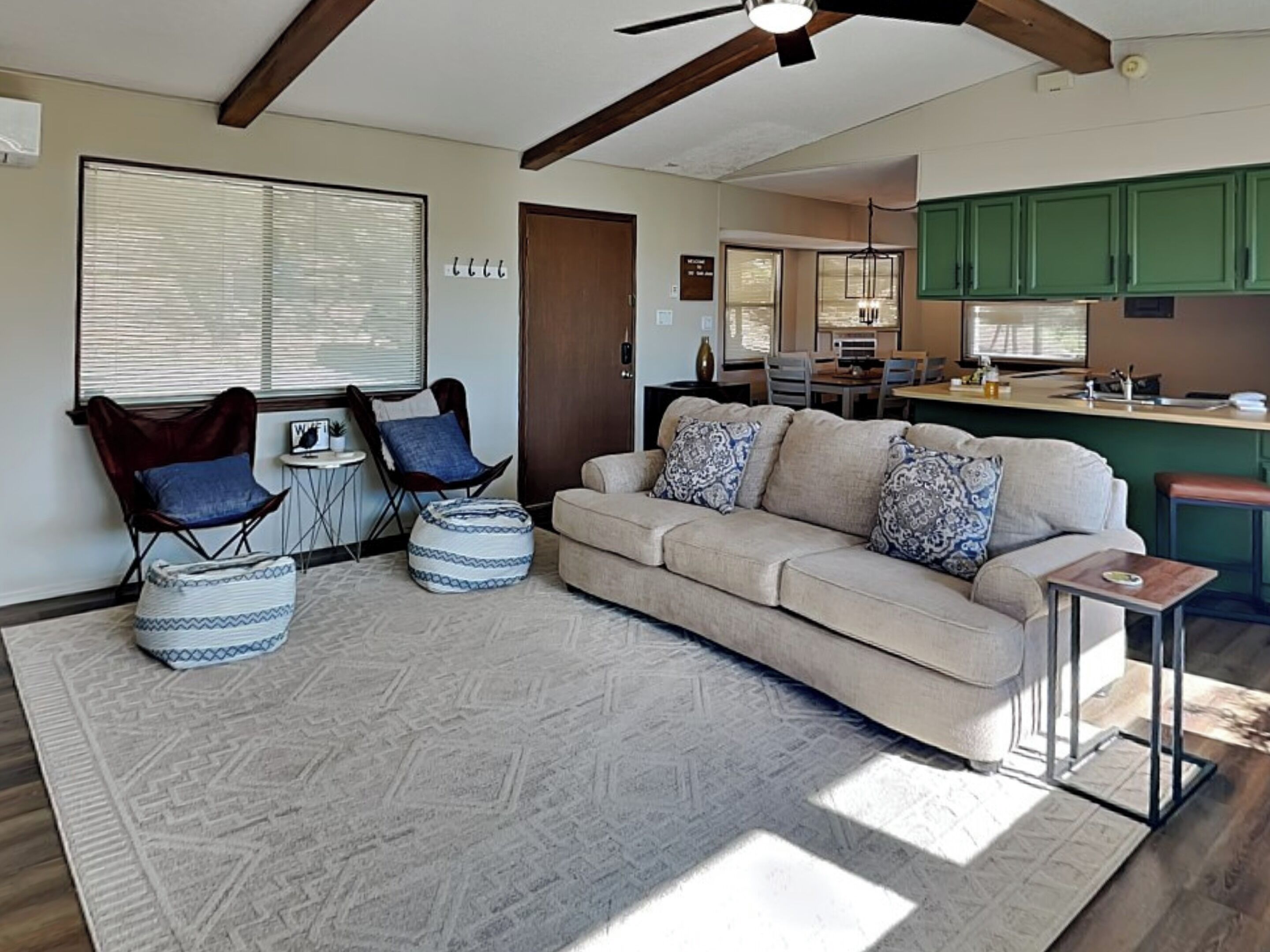 A living room with a beige sofa, patterned cushions, a coffee table, and a rug. There is a dining area with a table and chairs, and a kitchen with green cabinets.
