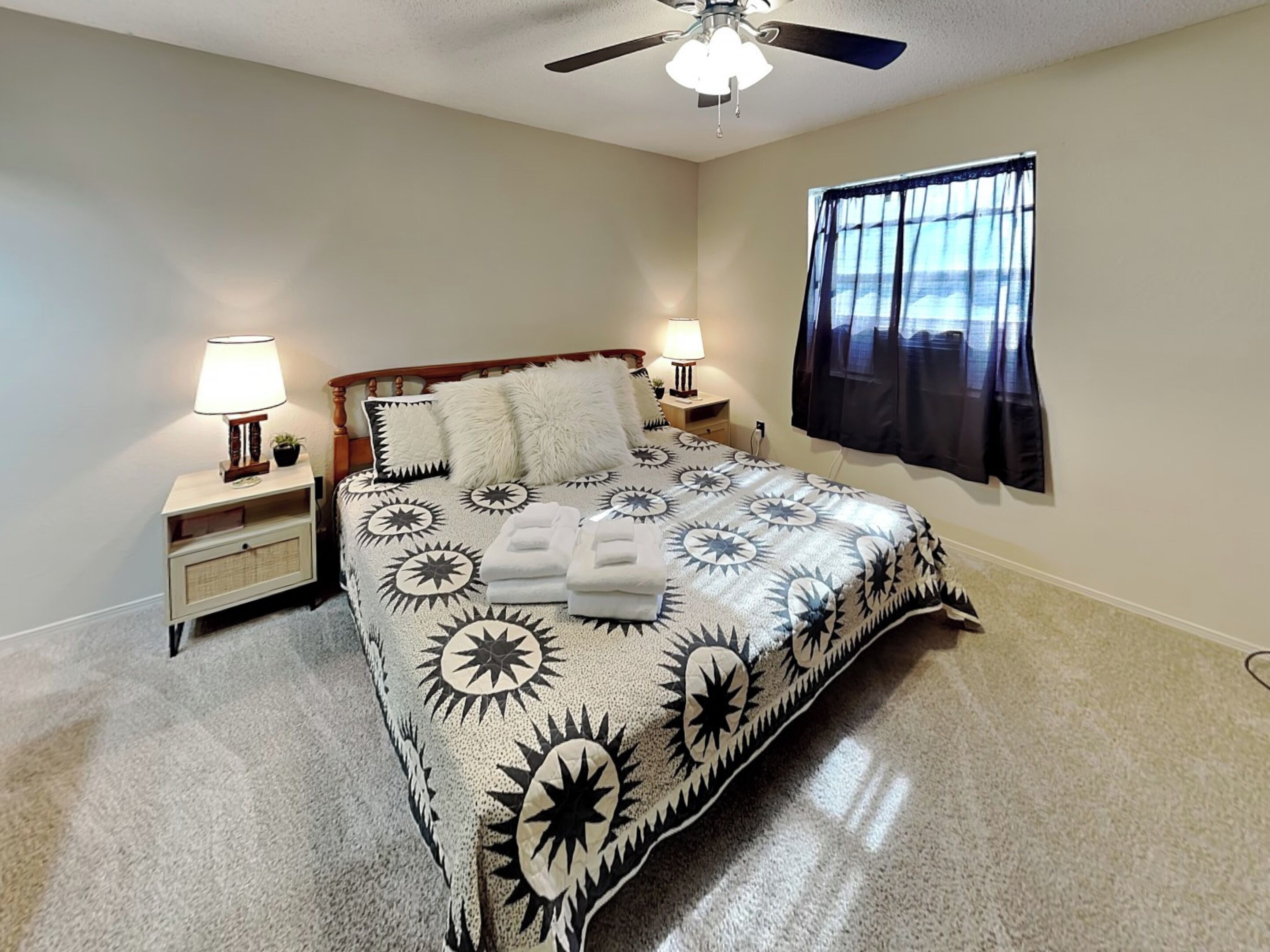 A bedroom with a bed, two bedside lamps, a ceiling fan, and a window with curtains.