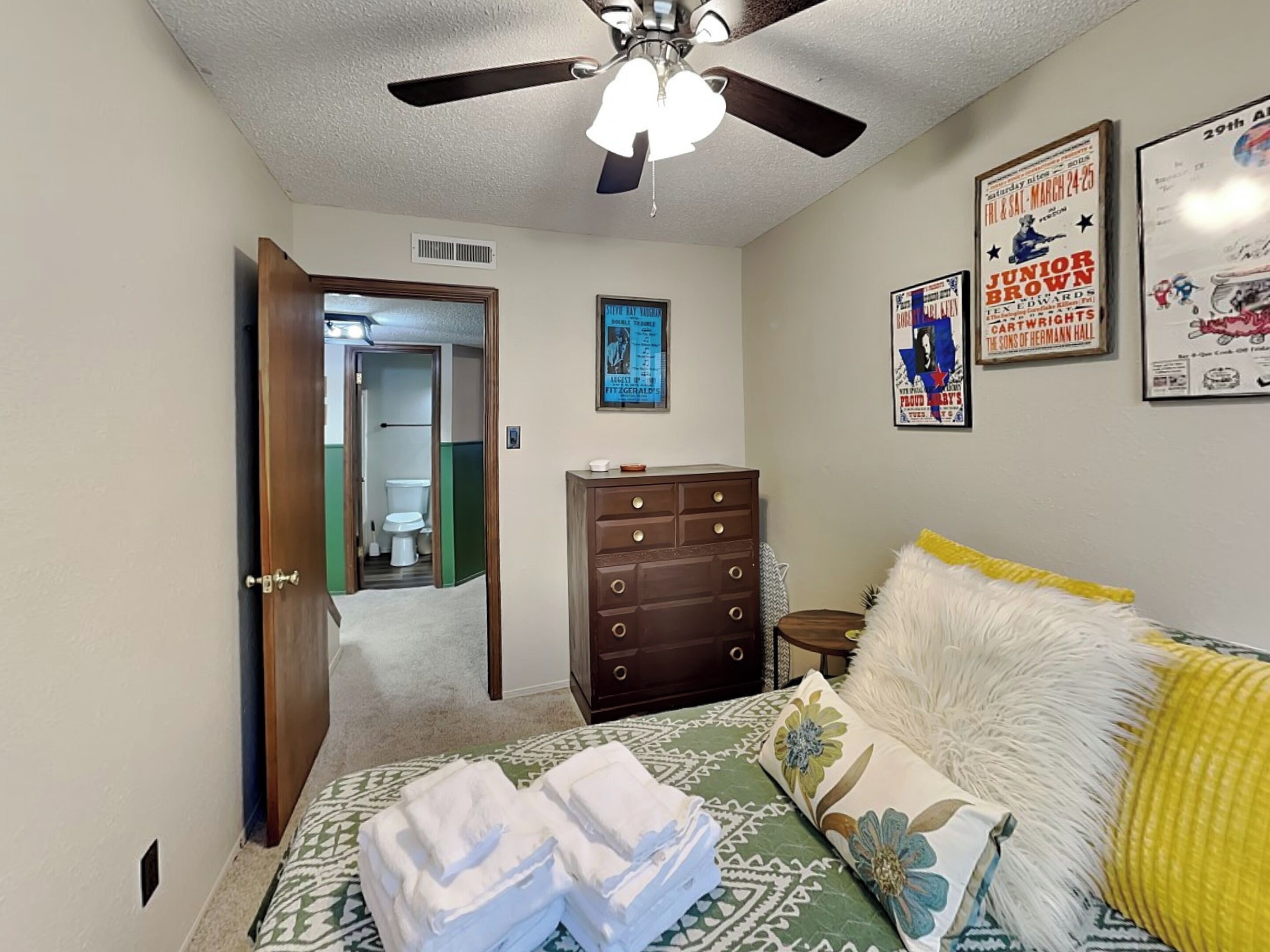 A bedroom with a bed, a dresser, a ceiling fan, and a door leading to a bathroom.