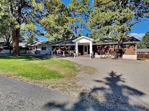A building with a covered porch, a grassy area in front, and trees surrounding the property. - Brooksland (Ruidoso)
