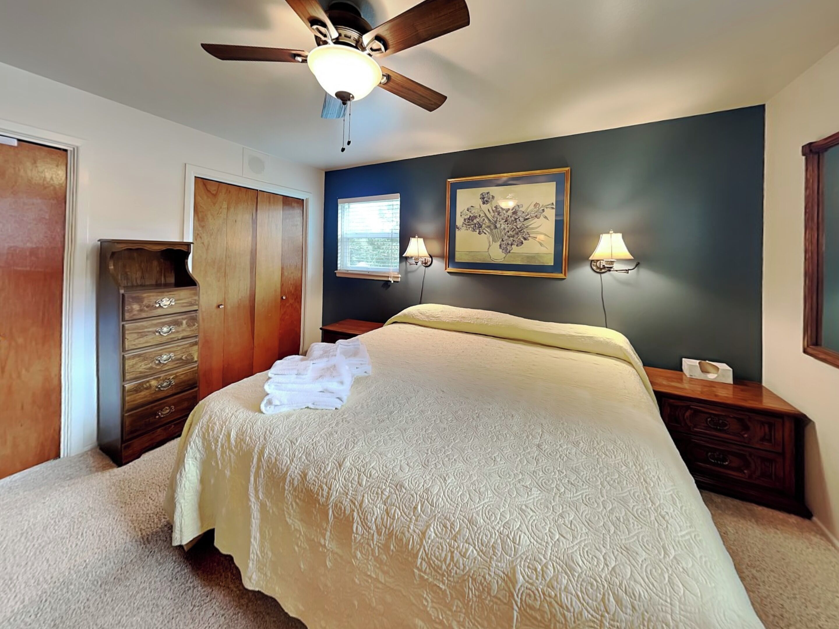 A bedroom with a bed, a ceiling fan, a dresser, a nightstand, a framed picture, and a window.