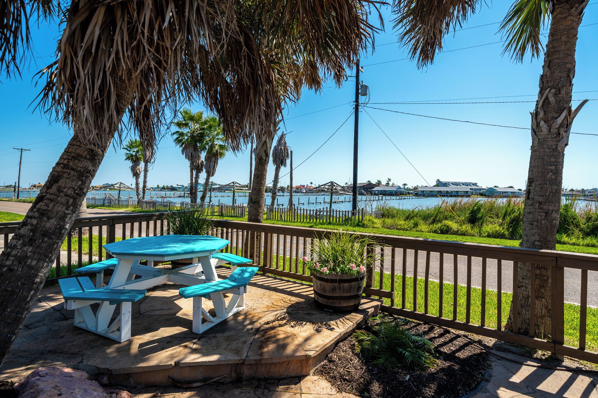 A picnic table with benches, surrounded by a wooden deck and a railing, overlooking a body of water with buildings in the background.