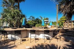 An outdoor bar area with a pool, surrounded by palm trees and a wooden fence.
