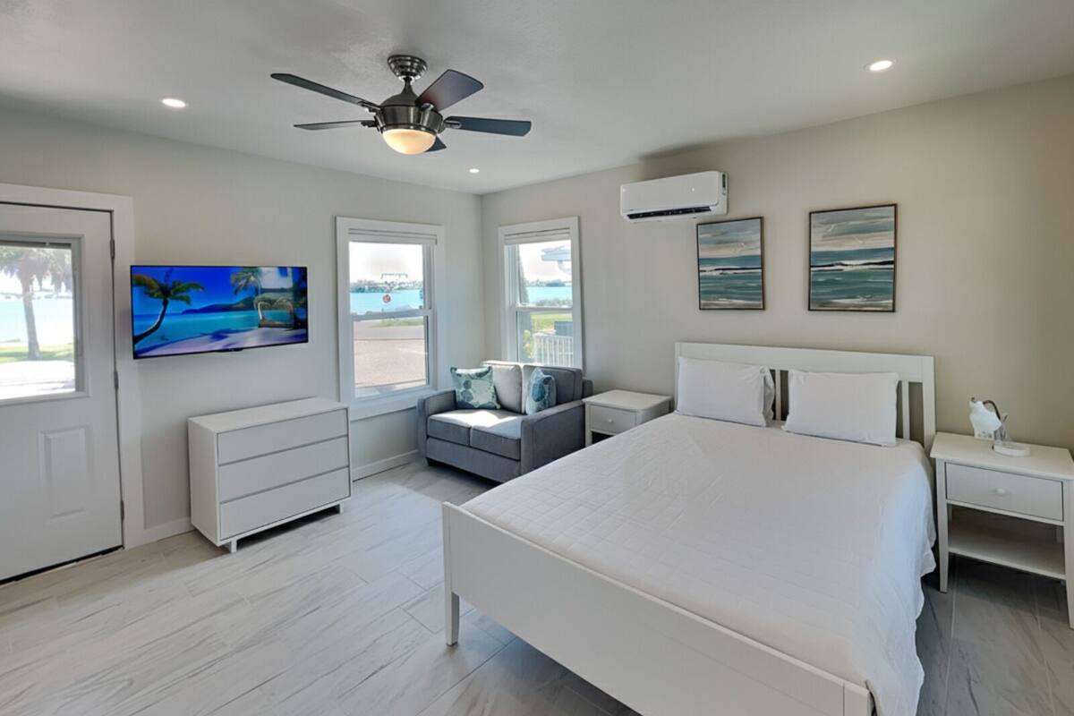 A bedroom with a large bed, a ceiling fan, a TV, and a window with a view of the outdoors.