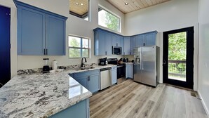 A modern kitchen with blue cabinets, granite countertops, a stainless steel refrigerator, and a large window offering a view of greenery. - Sunrise in the Cove (Sevierville)
