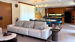 Ein modernes Wohnzimmer mit einer Couch, einem Esstisch und Stühlen. Es gibt eine Küchenzeile mit Holzschränken und einem Kühlschrank.