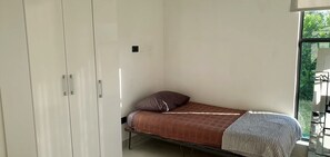 A single bed with a patterned blanket and a pillow, a wardrobe, and a window with a view of greenery.
