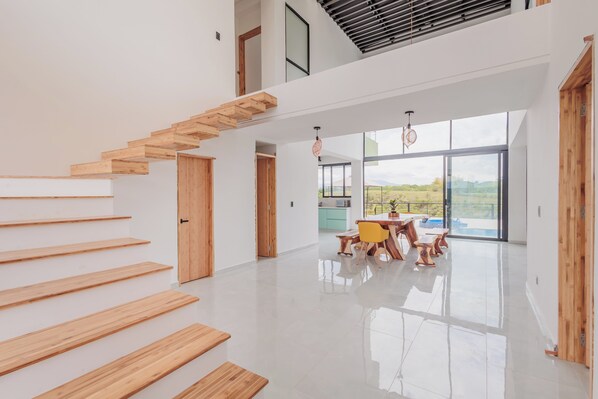 A spacious interior with a wooden staircase, a dining area with a table and chairs, and large windows offering a view.