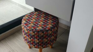 A small, round stool with a colorful geometric pattern, placed under a desk in a room with a tiled floor and a window.