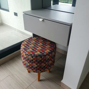 Un petit tabouret rond orné d’un motif géométrique coloré, placé sous un bureau dans une pièce dont le sol est recouvert de carreaux et qui possède une fenêtre.
