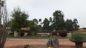 Zona ajardinada con un letrero que dice 'Sunrise Villas', con cactus, rocas y varios árboles.