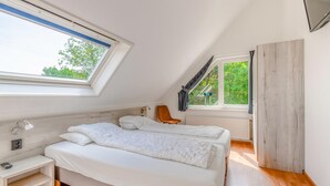 Ein Schlafzimmer mit schrägverdeckter Decke, einem Bett mit weißer Bettwäsche, einem hölzernen Nachttisch, einem Stuhl und einem Fenster mit Blick auf Grünflächen.