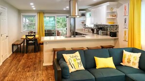 Een moderne keuken met een centraal eiland, een eethoek met een tafel en stoelen, en een woonkamer met een bank en kussens.