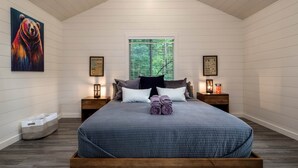 Une chambre avec un plafond en pente, un grand lit recouvert d’une courtepointe bleue, deux tables de chevet avec des lampes, une fenêtre donnant sur des arbres et un tableau représentant un ours accroché au mur.