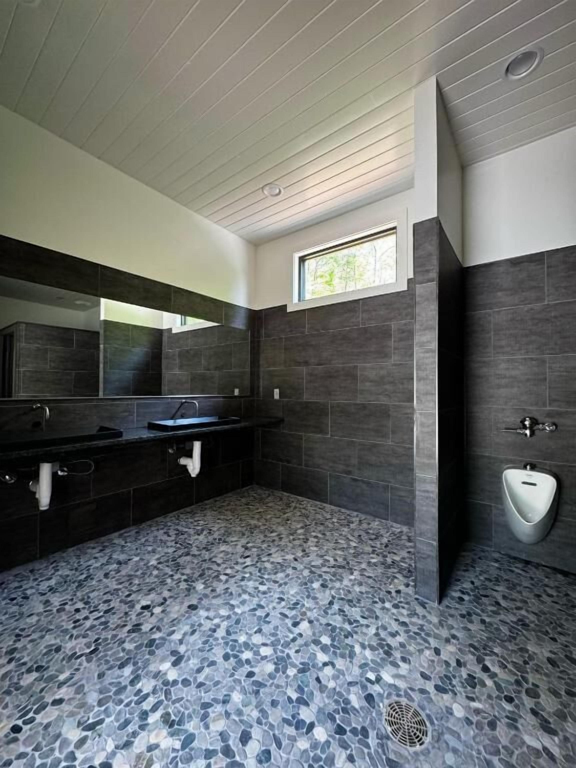 Ein modernes Badezimmer mit dunklen Fliesenwänden, einem Waschbecken aus Granit, einem großen Spiegel und einer Urinale.