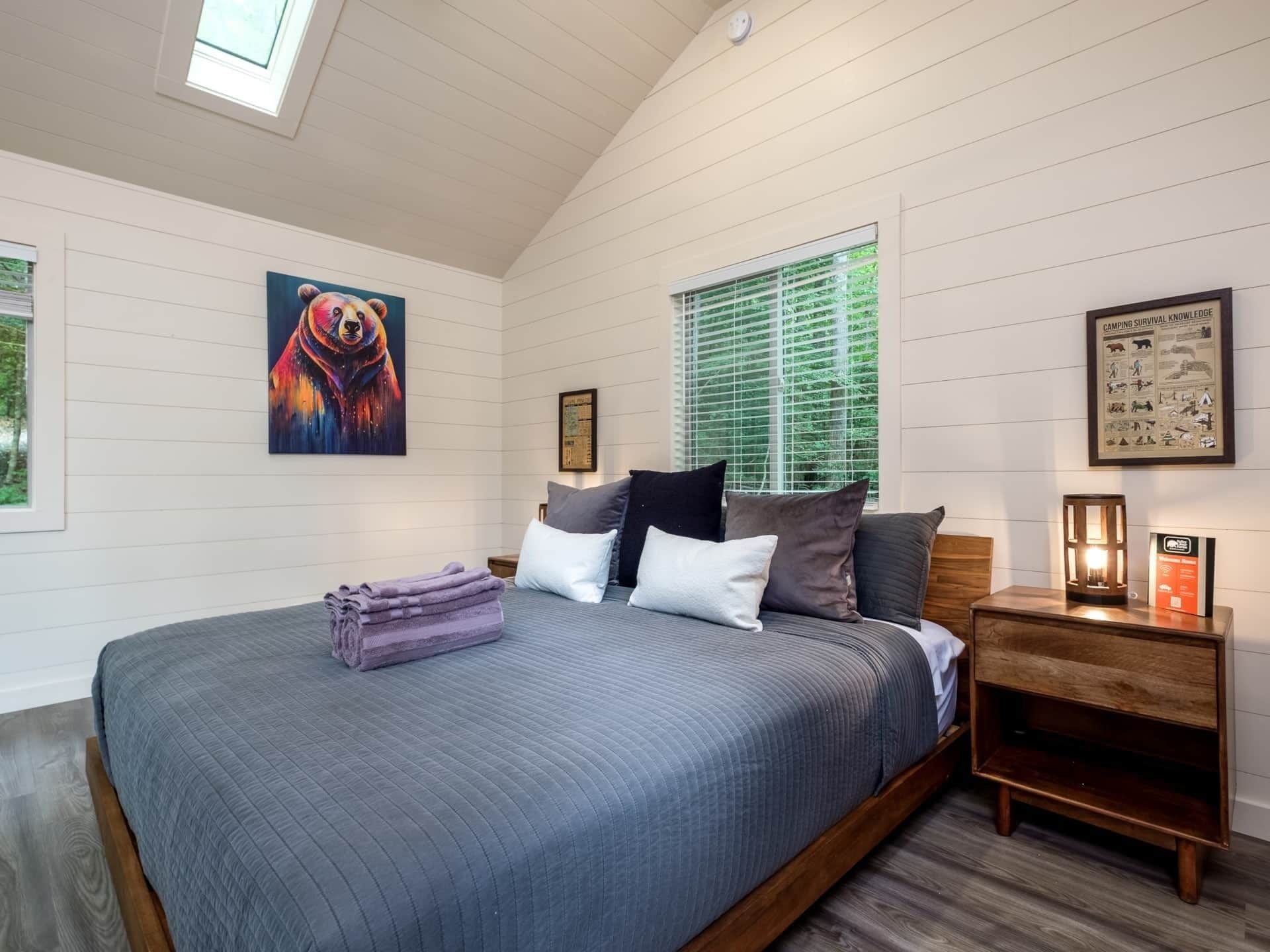 A bedroom with a large bed, a nightstand, a window with blinds, a painting, and a framed poster on the wall.