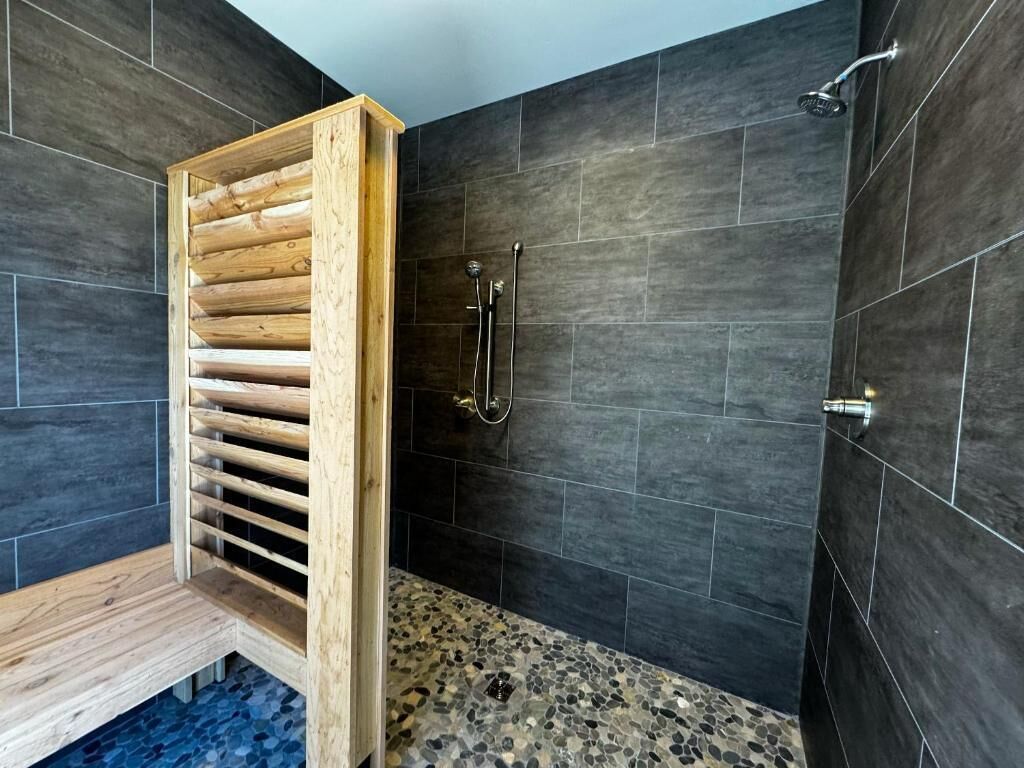 A modern bathroom with dark tiled walls, a wooden bench, and a shower area with a handheld showerhead.