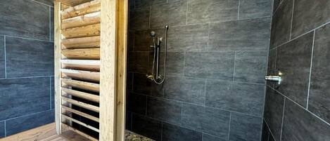 A modern bathroom with dark tiled walls, a wooden bench, and a shower area with a handheld showerhead.
