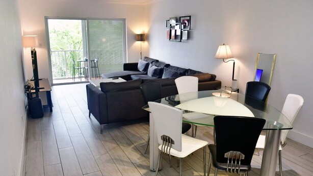 A modern living room with a glass dining table, white chairs, a dark sofa, and a balcony with a view. - Modern 2-bedroom apartment in North Miami Beach with AC (North Miami Beach)