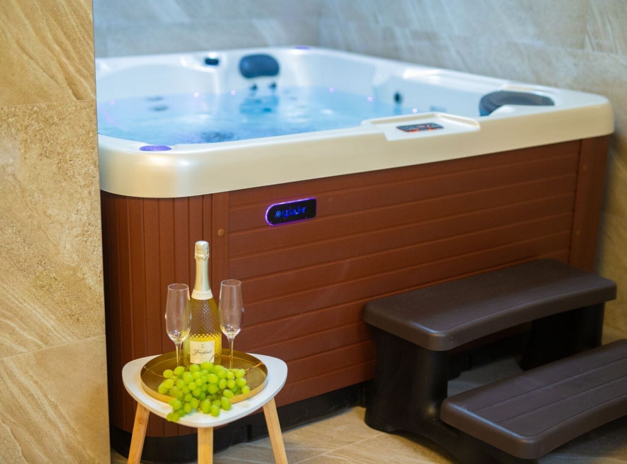 A hot tub with a control panel, a bottle of champagne, two glasses, and a tray of grapes.